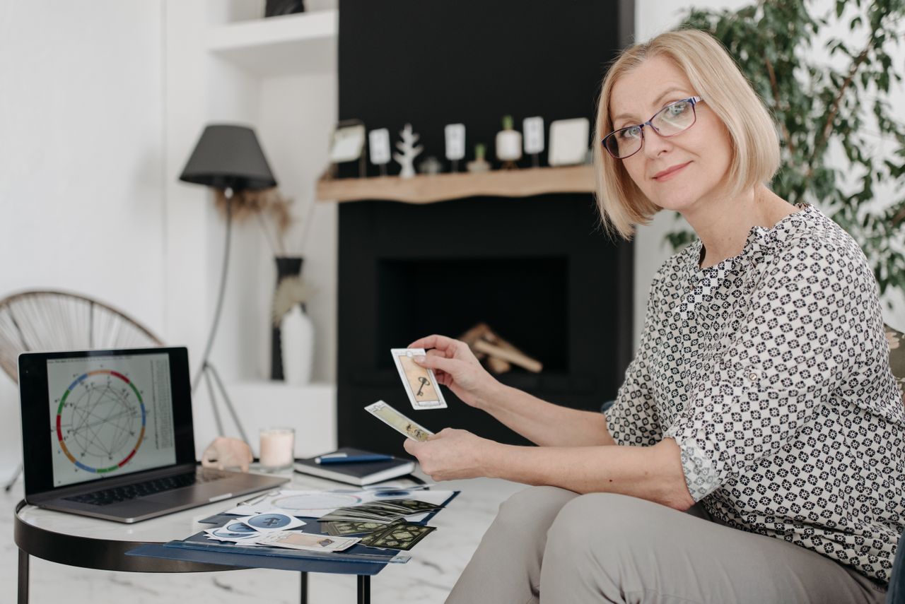 About Woman with tarot cards using laptop for astrology reading in cozy living room.